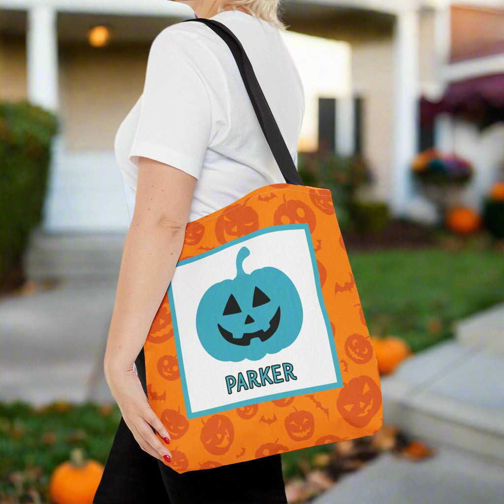 Person holding an orange Halloween tote bag with a teal pumpkin design and text, outdoors.