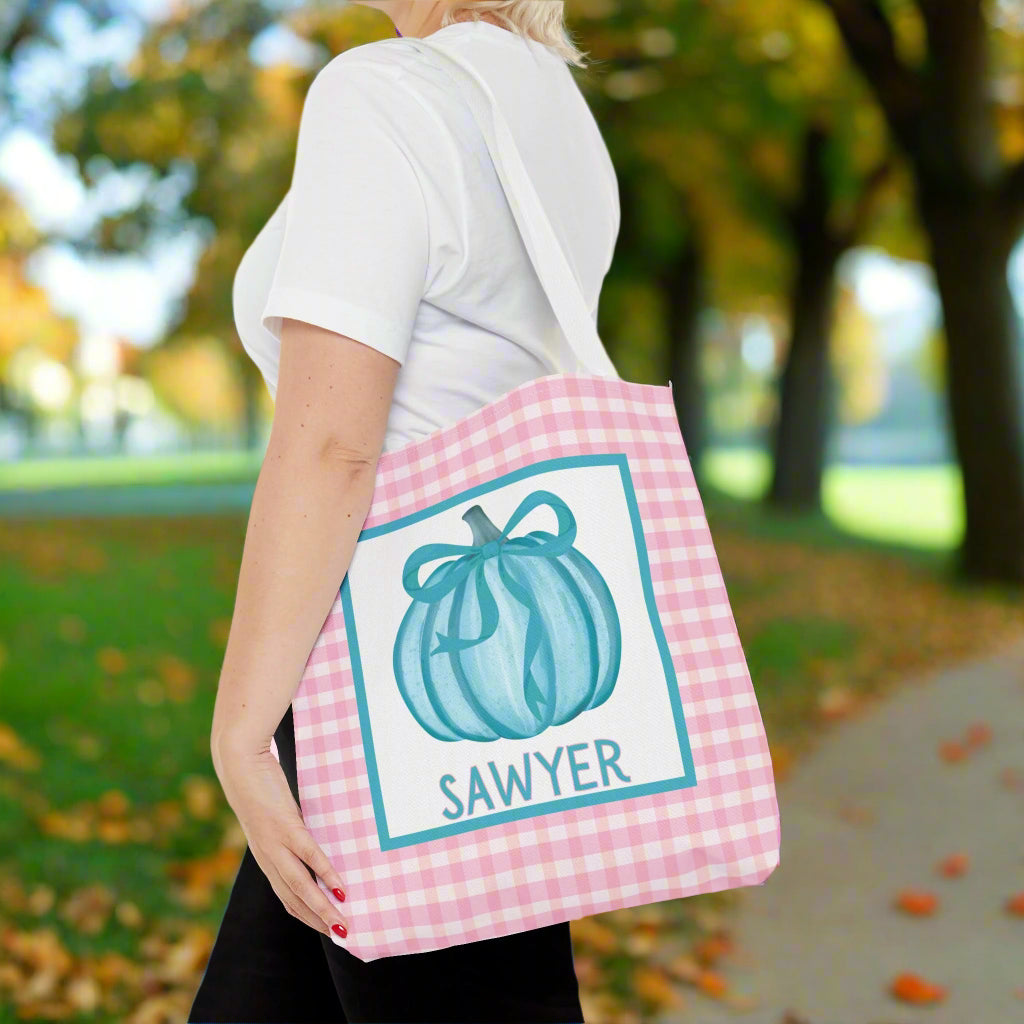 Person holding a tote bag with a teal bow pumpkin design and text on a pink check background.