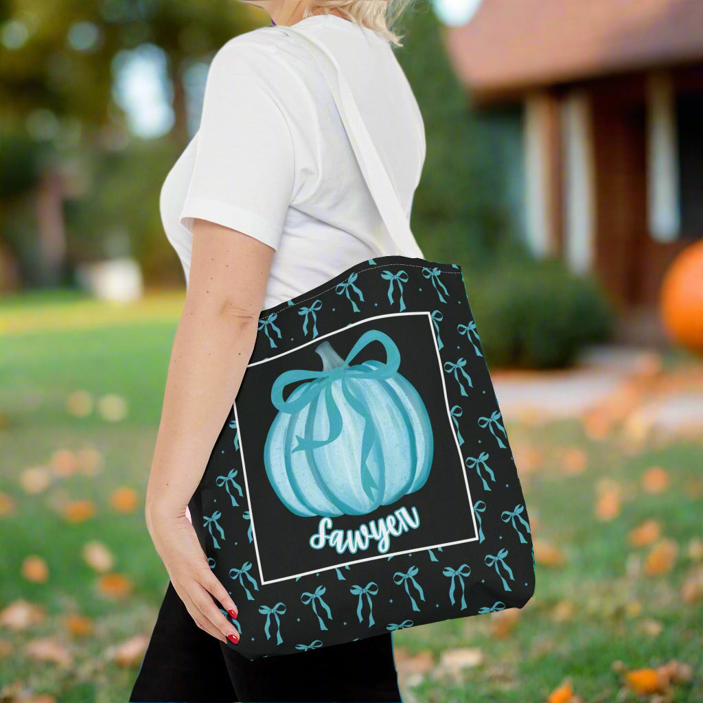 Person holding a tote bag with a pumpkin design and a name 'on a background with colorful teal bows.