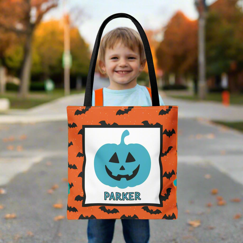 Child holding a Halloween-themed tote bag with a pumpkin design and a name on a blurred outdoor background.