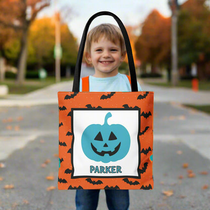 Child holding a Halloween-themed tote bag with a pumpkin design and a name on a blurred outdoor background.