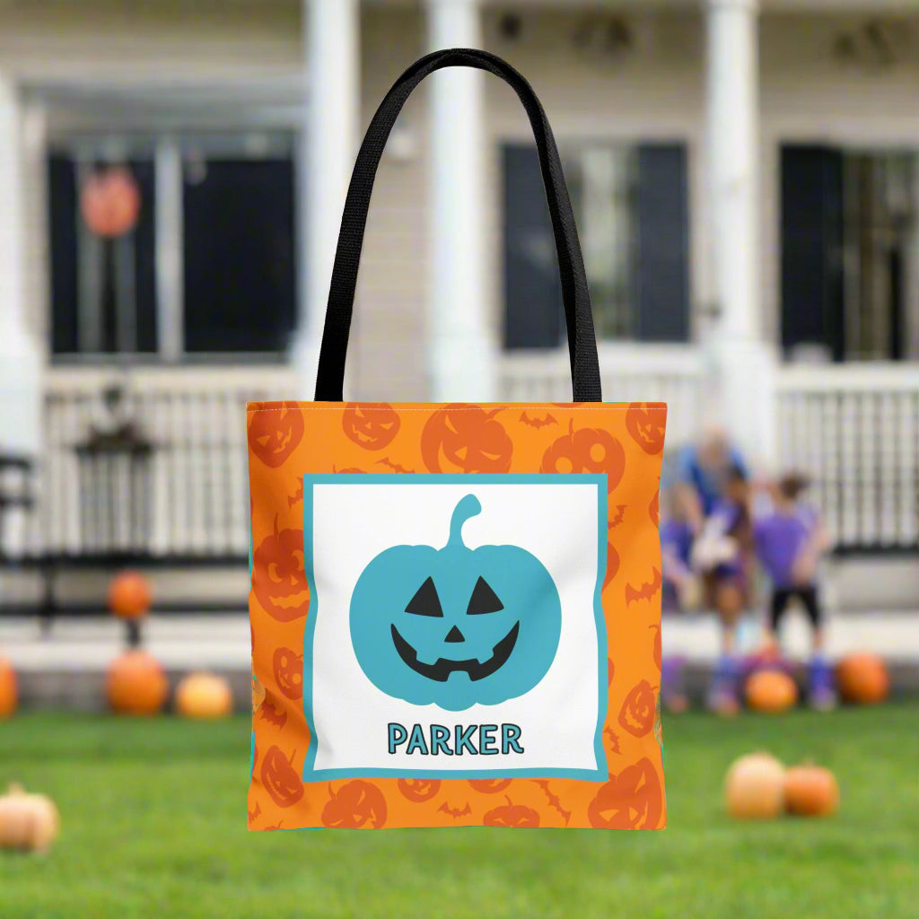 Orange tote bag with blue pumpkin design and text, set against a Halloween-themed background with pumpkins and a house.