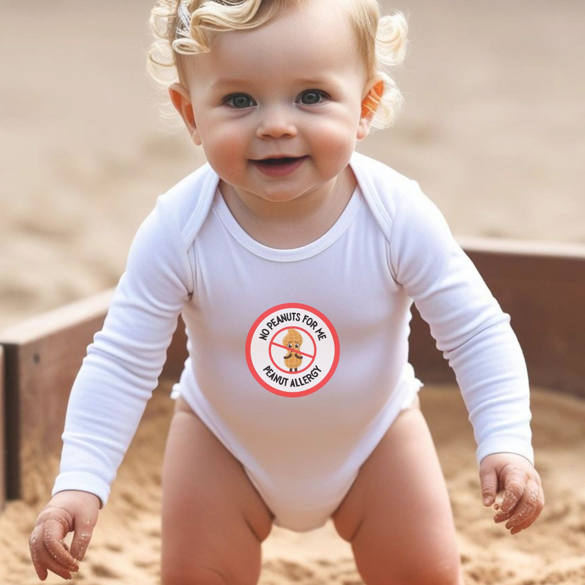 A baby wearing a white long sleeve onesie with a red logo that has text alerting to a peanut allergy. The onesie is being worn at a sandy location, possibly a playground.