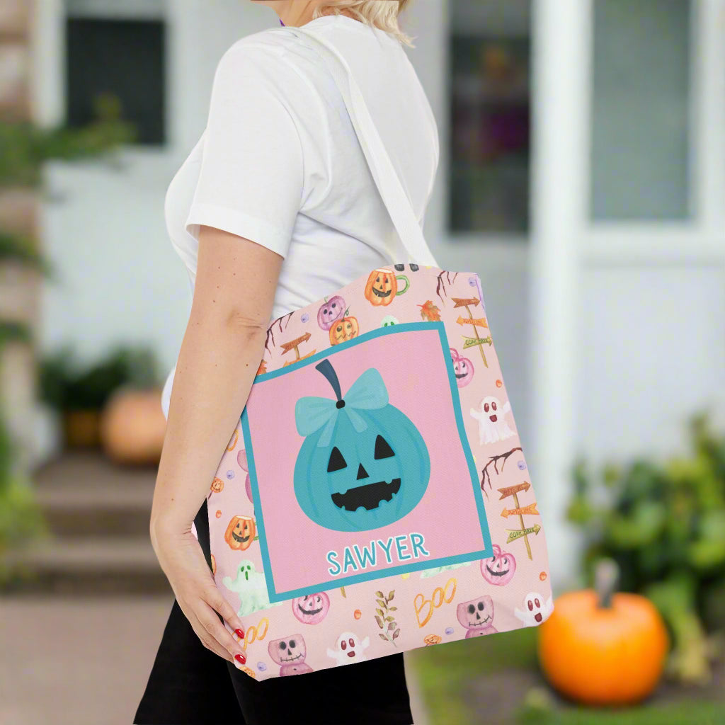 Person holding a Halloween-themed tote bag with a pink and purple pumpkin design on a pink background with teal bow pumpkin in middle.