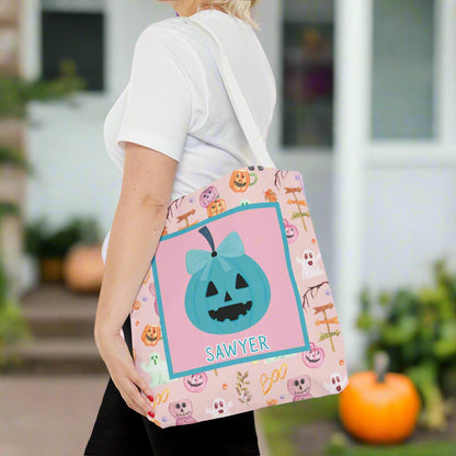 Person holding a Halloween-themed tote bag with a pink and purple pumpkin design on a pink background with teal bow pumpkin in middle.