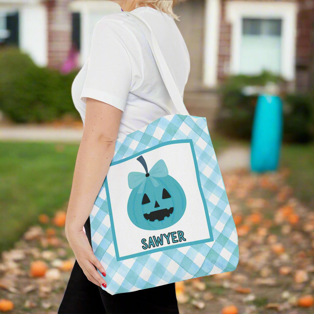 Person holding a tote bag with a pumpkin design and text, with a teal check background.